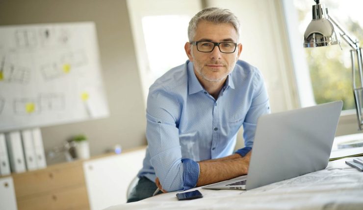 Photo d'un homme aux cheveux gris en chemise bleue accoudé sur une table avec un ordinateur et des documents