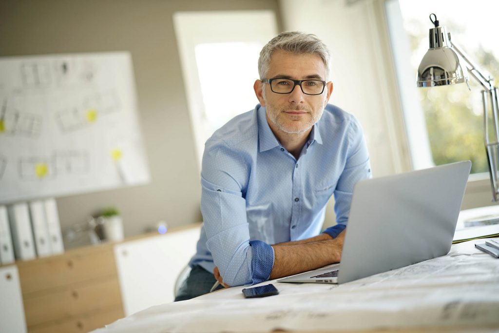 Photo d'un homme aux cheveux gris en chemise bleue accoudé sur une table avec un ordinateur et des documents